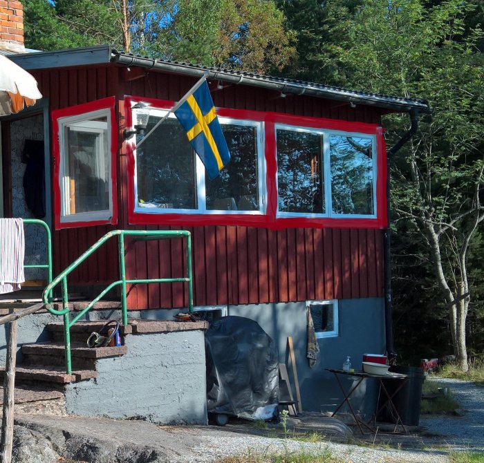 Husfasad med rödmålade fönsterramar utan spröjs och en svensk flagga, omgiven av natur.