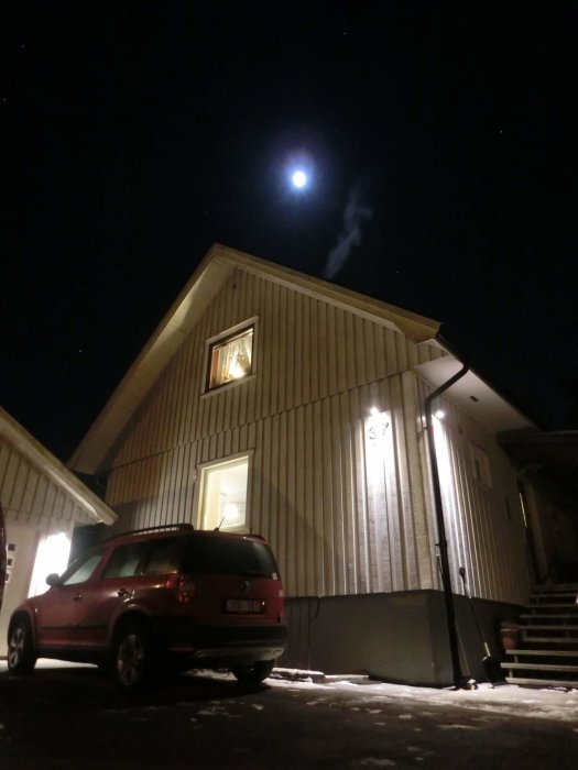 Husfasad på vintern med tända fönster, bil parkerad framför, och fullmåne på en mörk himmel.