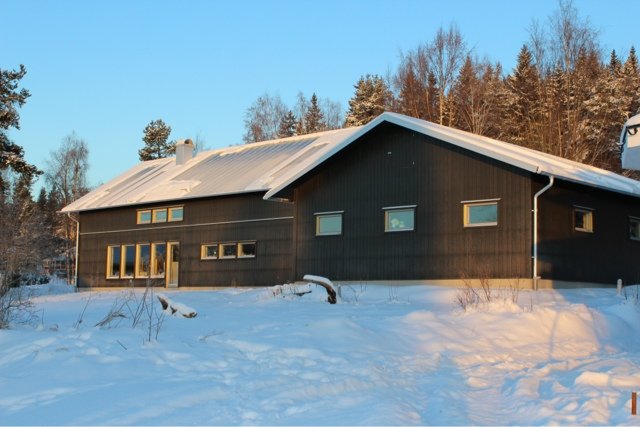 Mörkt trähus med snedtak täckt av snö, i ett snöigt landskap med träd i bakgrunden under en klar himmel.