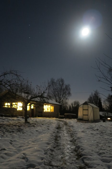 Nattvy med fullmåne över ett snötäckt landskap, fotspår och hus med tända fönster.