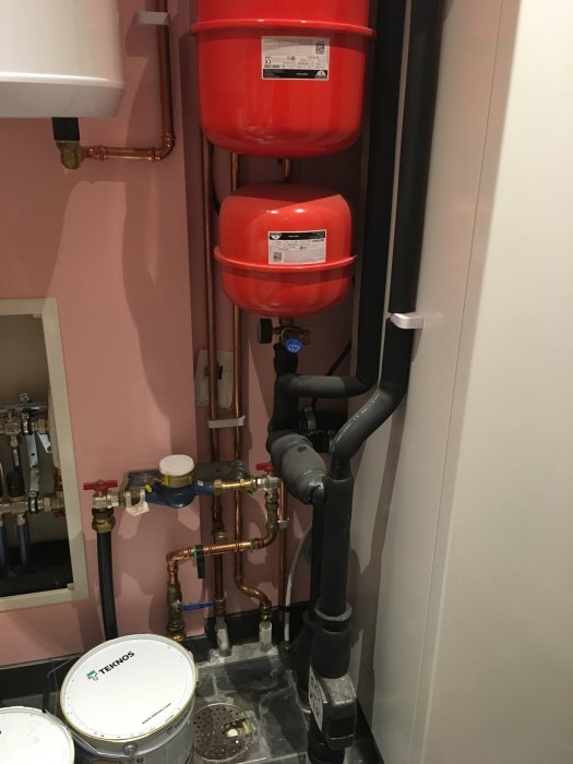 Installation av två röda expansionskärl ovanför koppar- och svarta rörledningar vid en vägg.