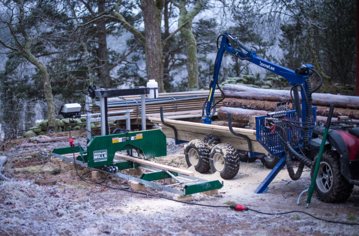 Portabelt sågverk utomhus med stockar, ansluten kran och fyrhjuling i skogsmiljö.