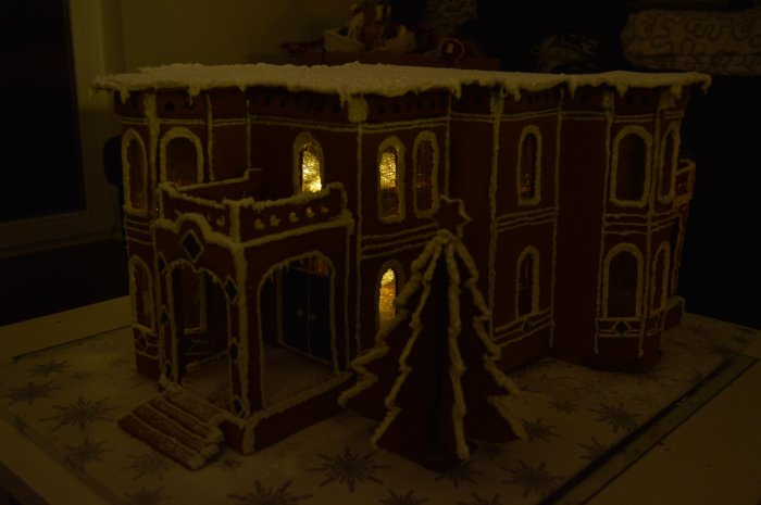 Pepparkakshus inspirerat av viktoriansk italieniserande stil med tända fönster och snö på taket, vid sidan en pepparkakstomte.