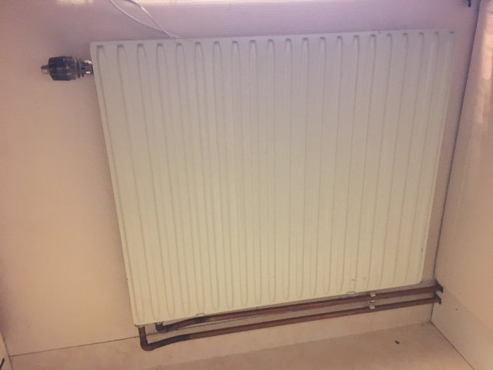 Vit väggmonterad radiator, kopplad till rör, svagt upplyst rum, hörninstallation.