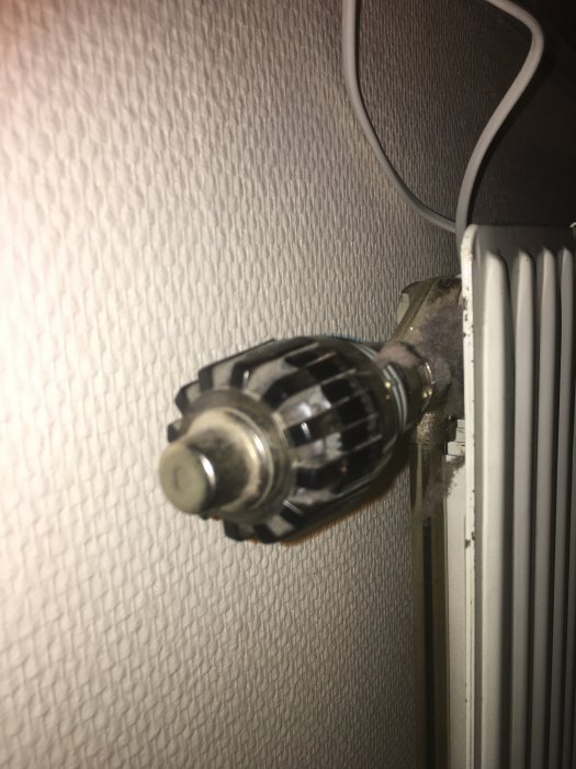 Termostatventil på radiator utan ratt mot en vit tapetserad vägg.