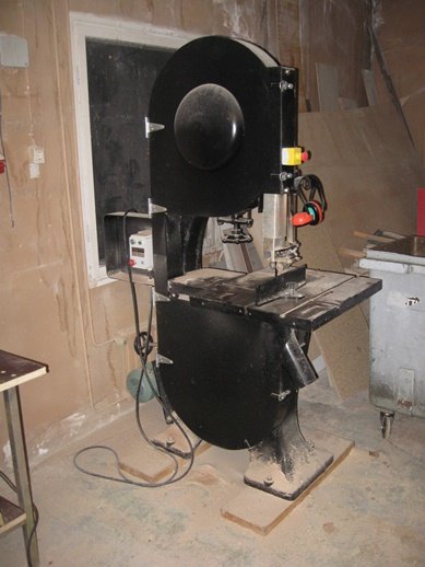 Stor bordsbandsåg i en verkstad med oavslutade väggar och byggmaterial i bakgrunden.