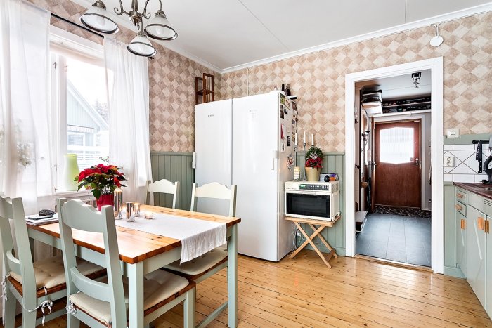Ljust kök med trägolv, pastellgröna skåp, vitt kylskåp, retro tapet och matbord med blommor.