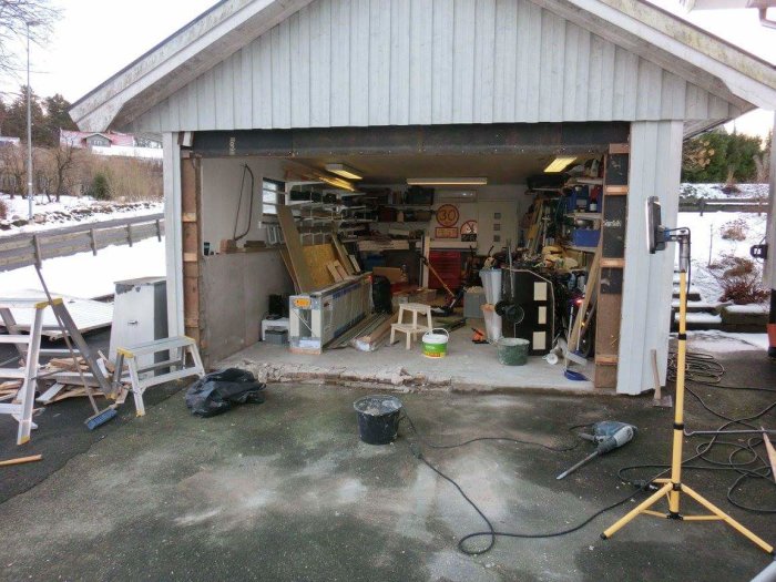 Garage under ombyggnad med röriga verktyg och material, dörr och vägg delvis rivna.