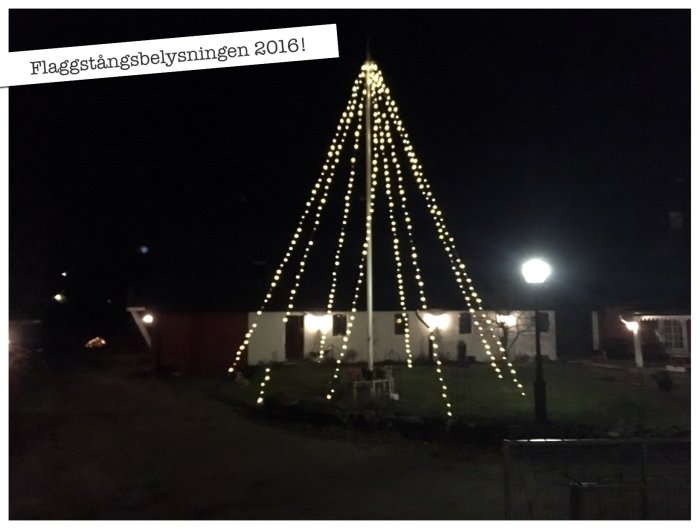 Julbelysning med ljusslingor i form av en julgran runt en flaggstång i en trädgård på kvällstid.