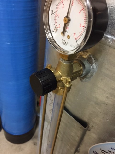 Manometer med två vred vid ett nivåglas på en vägg, med en blå rörledning i bakgrunden.