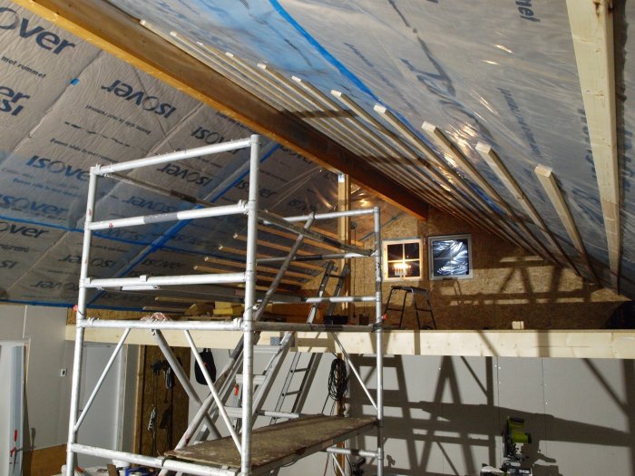 Byggarbetsplats med otätat tak och glespanel, byggställning, isolering och verktyg synliga.