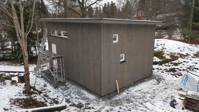 Nybyggt hus i vinterscen med träpanel, snötäckt mark och stege lutad mot väggen.