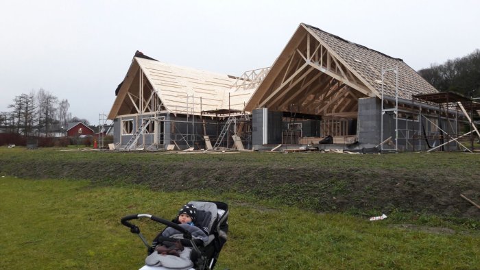 Barn i barnvagn framför pågående byggnation av trähus med öppen takstomme.