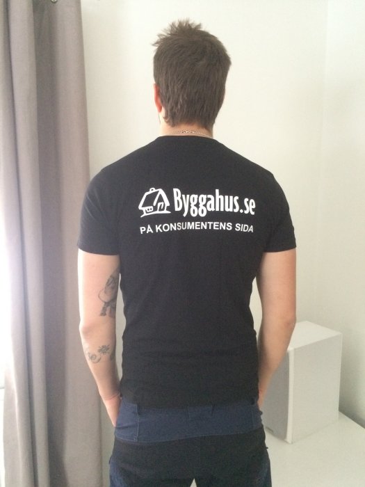Man i svart t-shirt med logga "Byggahus.se" synlig från ryggen, tatuerad arm syns delvis.