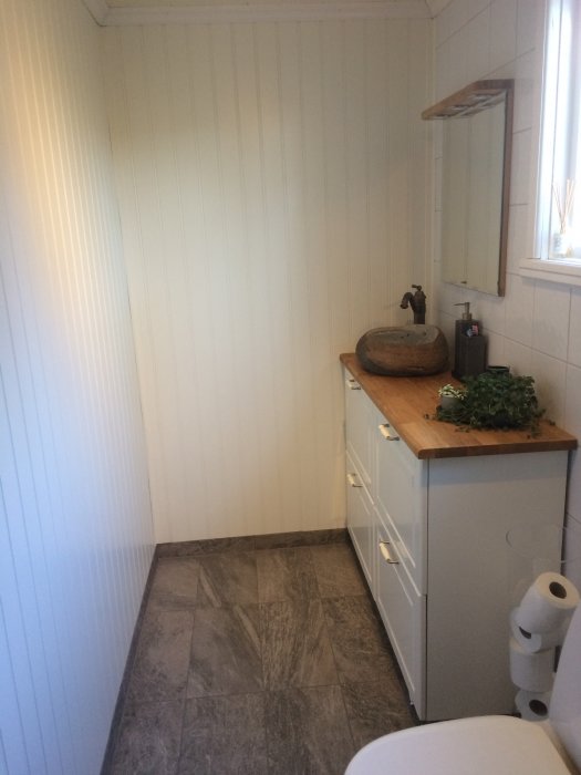 Nyrenoverat badrum med vita väggar, grå skåp, träbänkskiva och stengolv.