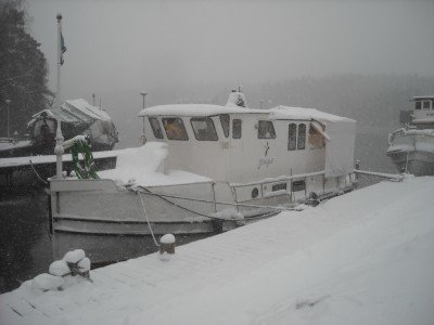 Husbåt täckt av snö förtöjd vid en brygga under en snöig vinterdag.