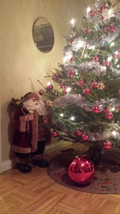 Tomtefigur bredvid en julgran dekorerad med röda kulor och glitter i ett vardagsrum.