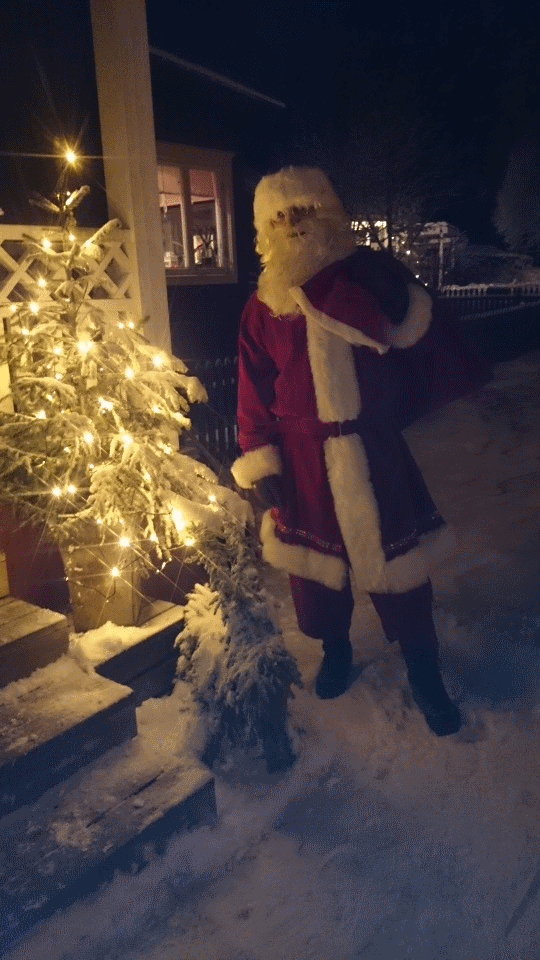 Person utklädd till jultomten står bredvid ett ljusdekorerat träd i snöigt landskap på kvällen.