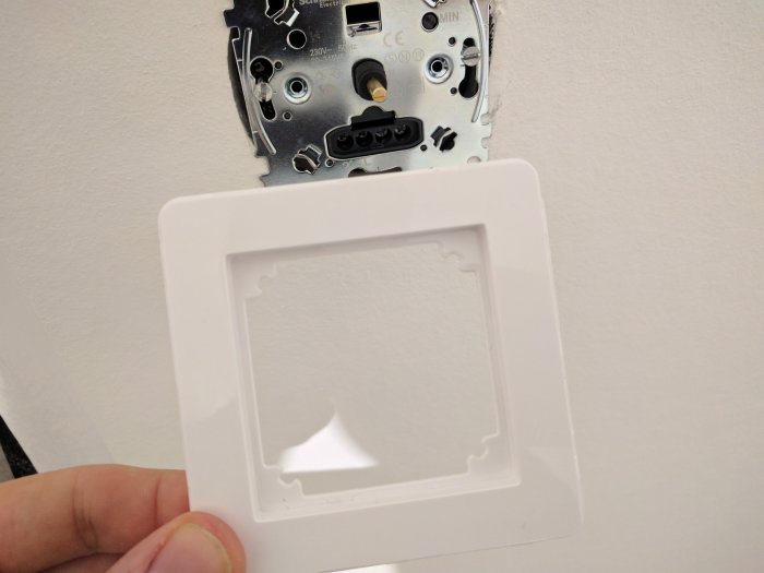 Hand håller ett löst täcklock framför en väggmonterad dimmer från Schneider.