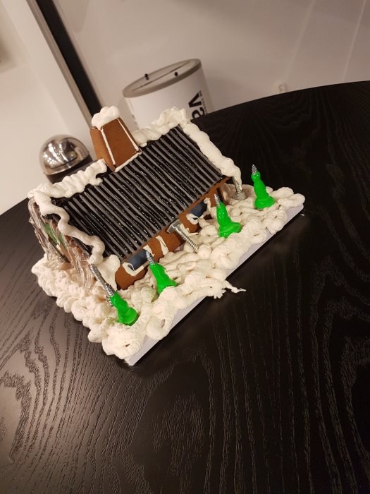 Humoristisk pepparkakshusmodell med vitt skum som tak och fog, gröna figurer och svarta linjer, på mörkt bord.