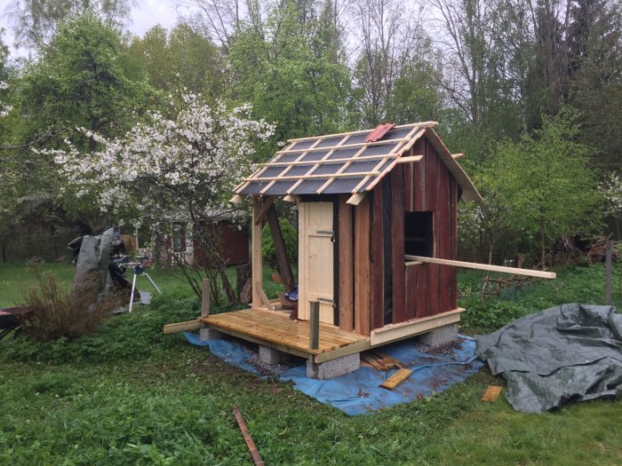 Hemsnickrad stuga med återanvänd panel och solcellspaneler på taket framför blommande körsbärsträd.