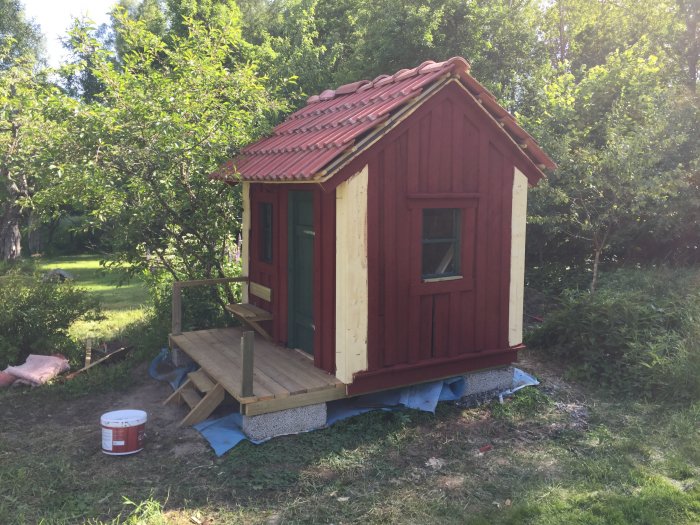 Nybyggt rött lekstuga med gröna detaljer och plexiglasfönster, trätrall och burk med färg utomhus.