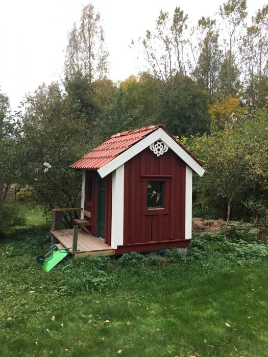 Nyrenoverat rött lekstuga med grön dörr, plexiglasfönster och dekorativ snickarglädje i trägården.