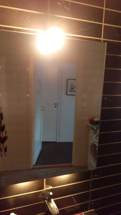 Spegelbild av ett mörkt kaklat badrum med syn av en korridor genom spegelreflektionen.