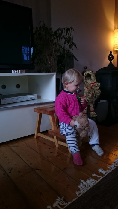 Ett barn sitter på en hemmagjord pall av björk, leker med en nalle och en figur.