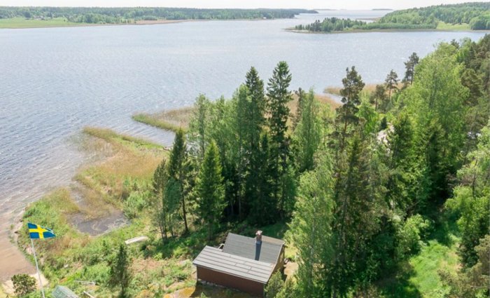 Flygfoto av en liten stuga vid Vänern nära skog och vatten, med en svensk flagga i förgrunden.