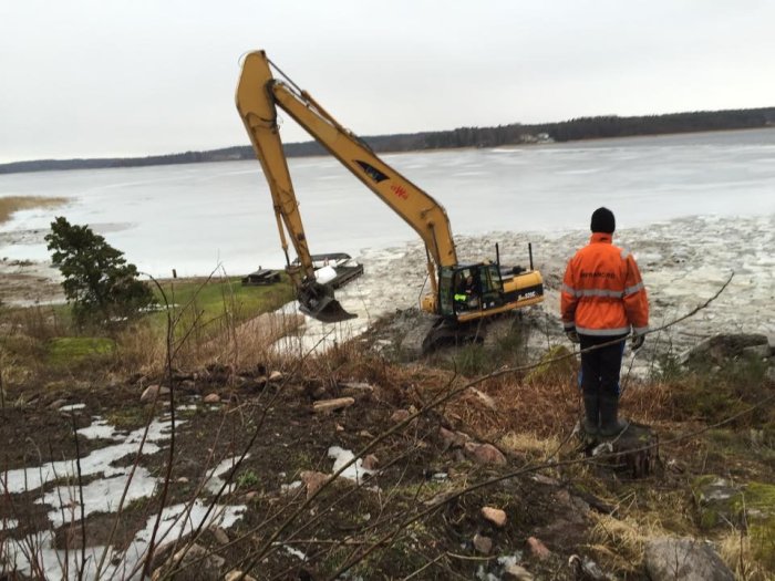 Man i orange jacka som övervakar en grävmaskin vid en isbelagd sjö under röjningsarbete vintertid.