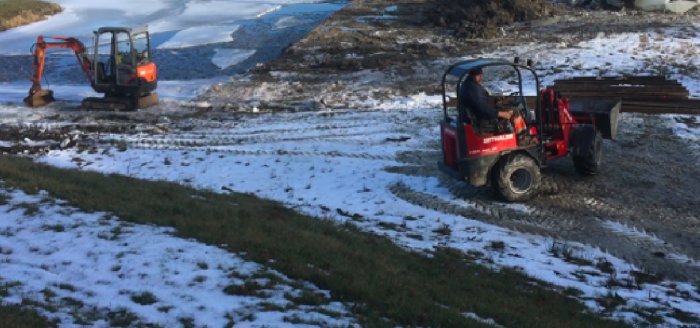 Röjningsarbete vid en strand med snö, en grävmaskin och ett fordon som drar timmer.