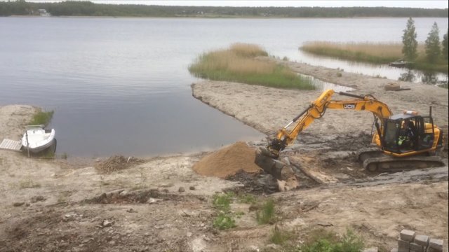 Grävmaskin vid arbete på strandkanten med utsikt över en sjö, sandhögar och en liten båt vid kajen.