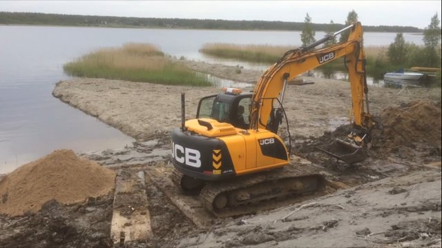 Grävmaskin av märket JCB arbetar med markförberedelse vid en strandkant, med sjö och vass i bakgrunden.