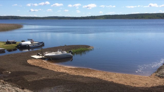 Motorbåt vid en brygga längs en strandkant med utsikt över en sjö under klar himmel.