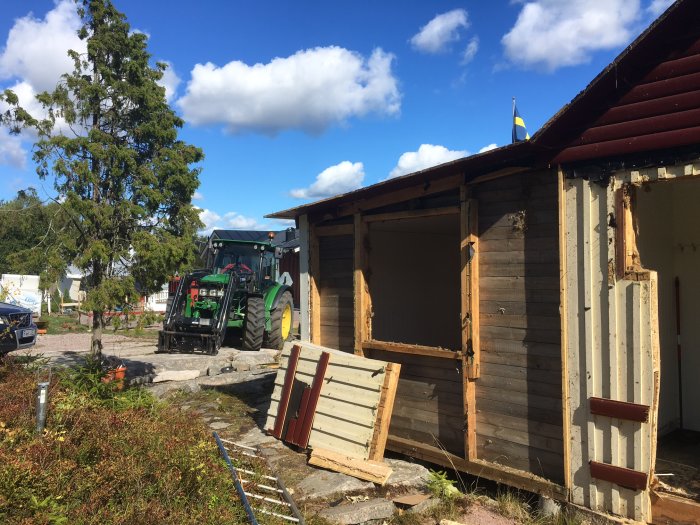 Delvis nedmonterad stuga med synliga väggstommar och en traktor i bakgrunden.