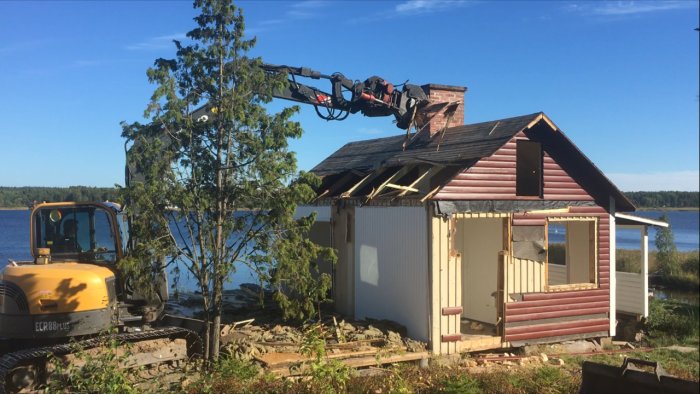 Grävmaskin river taket på en gammal stuga vid en sjö.