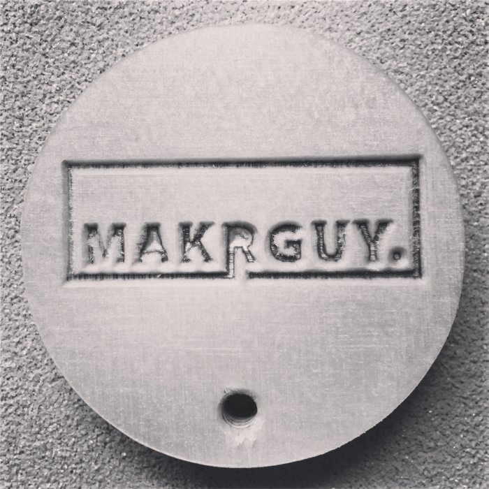 Nyckelbricka fräst i metall med texten "MAKRGUY" inuti en rektangel, avslutat CNC-projekt.