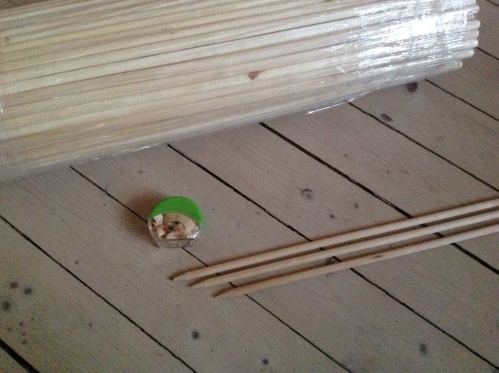 Oanvända blompinnar bredvid vässade pinnar och en grön pennvässare på trägolv.