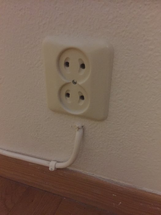 Ojordat vägguttag med en vit kabel som går ut genom ett hål i väggen vid golvlisten.