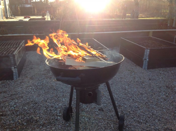 En grill med flammande vedbitar i en trädgård mot en solnedgång.