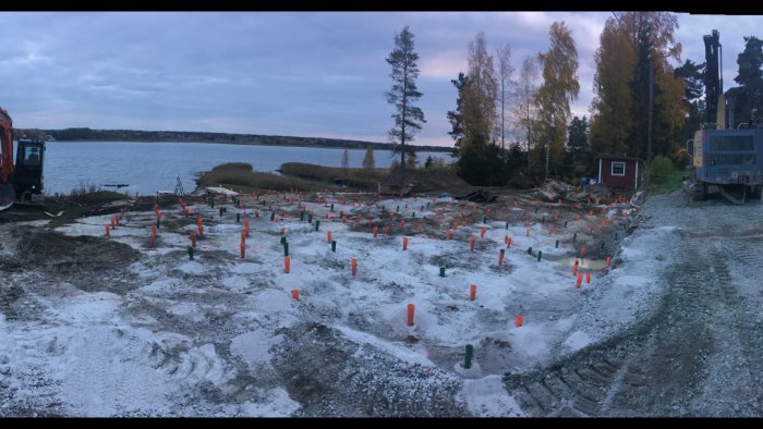 Byggarbetsplats vid vatten med markeringspinnar, grävmaskiner och ojämn terräng under skymning.