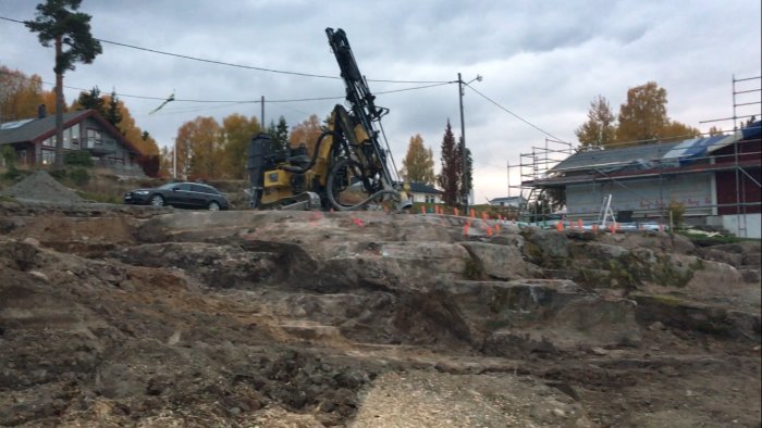 Byggarbetsplats med borrningsrigg, uppriven mark, byggnadsställning och hus i bakgrunden. Träd med höstfärger syns.