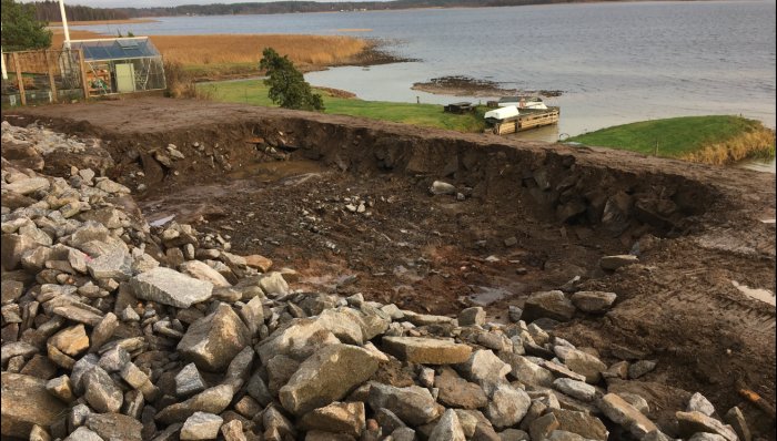 Markarbete vid sjö med stora sprängstenar och avskalat berg efter demolering.