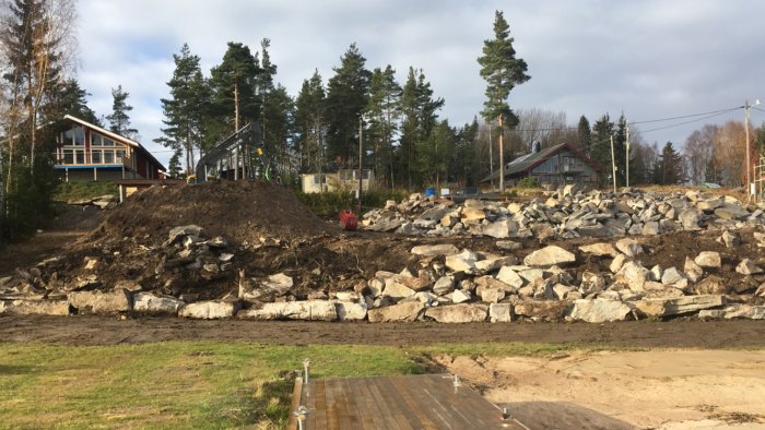 Markarbete vid sjö med sprängsten och grävmaskin bredvid hus och skog.