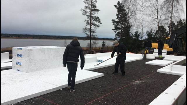 Arbetare förbereder isolering för gjutning av grund vid bygge, med markarbeten synliga och sjö i bakgrunden.