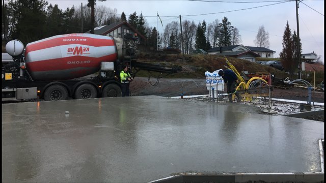 Arbetsplats där en betonggrund gjuts med en cementbil och arbetare under lätt regn.