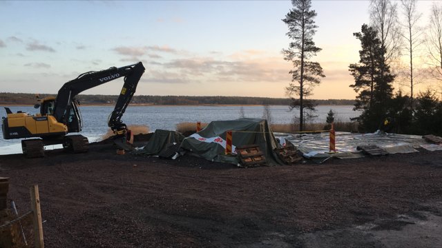 Nygrundad byggarbetsplats med isolerade presenningar och grävmaskin vid sjökant i gryning.