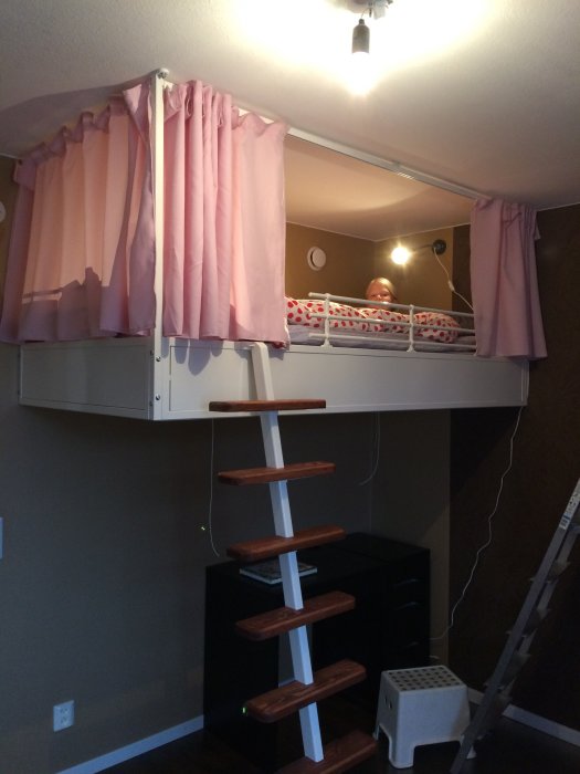 Loftsäng monterad mot vägg med rosa gardiner, stege, och synligt barn som ligger i sängen.