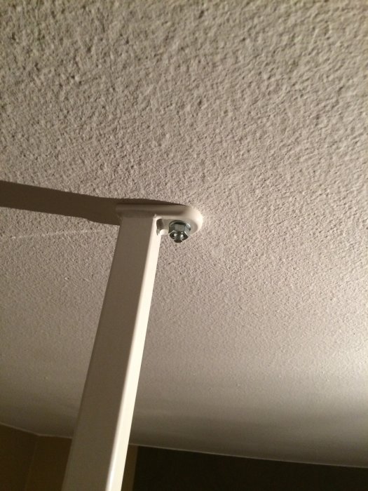 Installation av en vit gardinstång fästad mot en texturerad vit takyta med skruv och beslag.